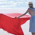 female laying down red blanket with fringe onto in the sand on the beach near the ocean