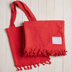 red beach blanket with fringe and a matching bag on a white wood background