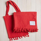 red beach blanket with fringe and a matching bag on a white wood background