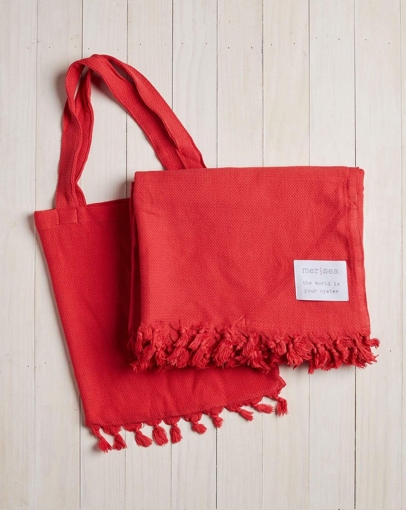 red beach blanket with fringe and a matching bag on a white wood background