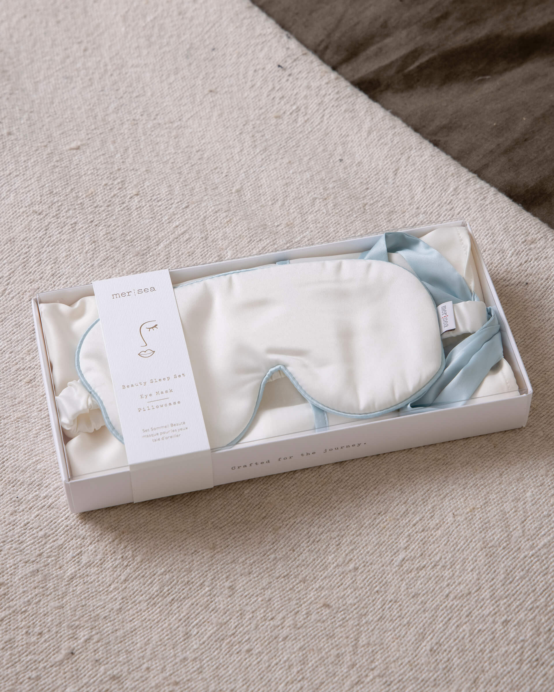 white satin sleep mask with light blue strap in a white box sitting on a neutral carpet
