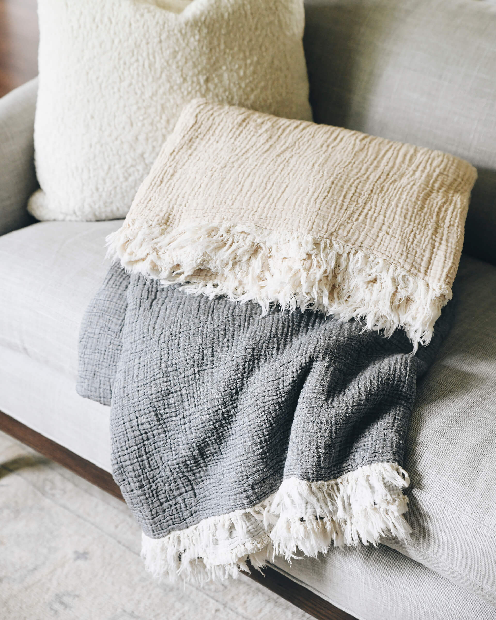 grey and light beige gauze blanket with white fringe detailing laying folded stacked on couch