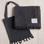dark grey beach blanket with fringe and a matching bag on a white wood background