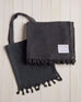 dark grey beach blanket with fringe and a matching bag on a white wood background