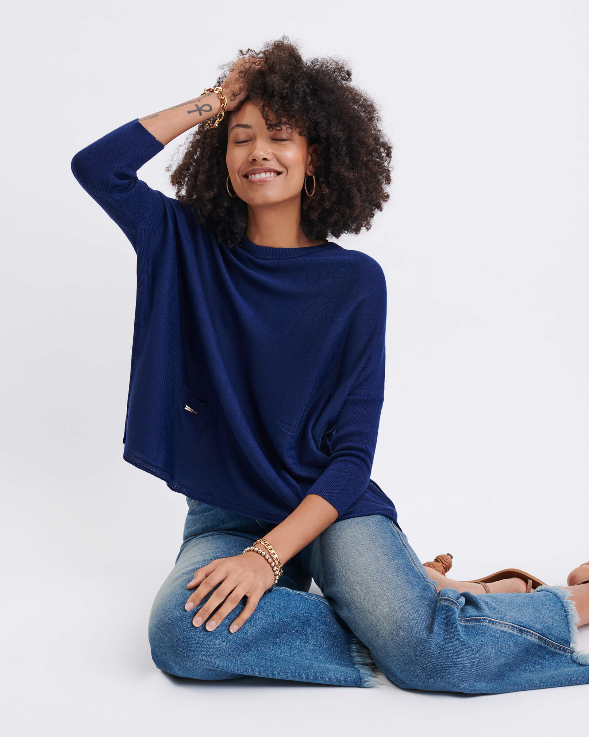 curly haired female wearing dark blue sweater sitting with a hand to her head on a white background