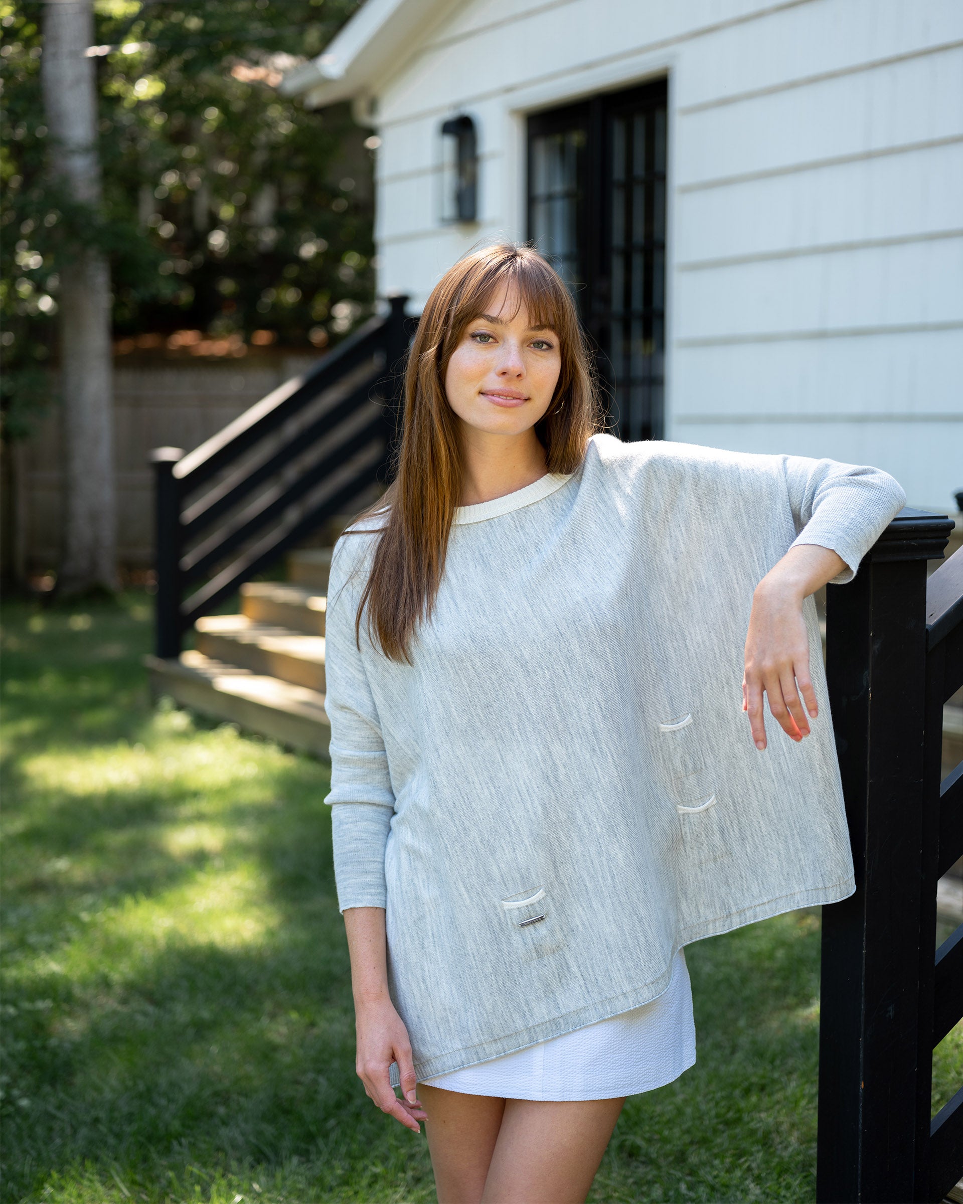 brunette female wearing grey and white sweater standing outside leaning against a railing on a porch