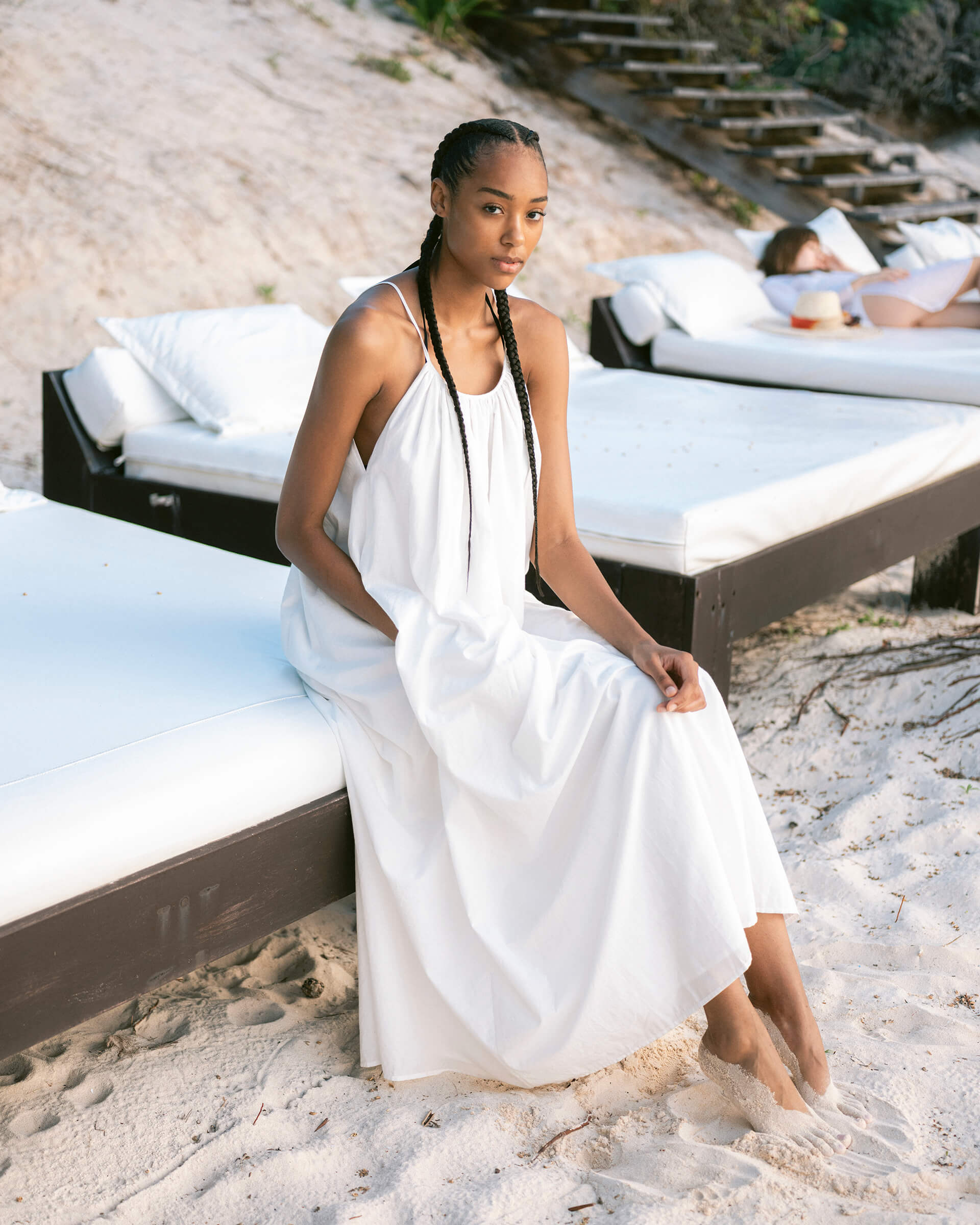 female wearing white maxi dress with a hand in pocket sitting on a day bed on the beach 