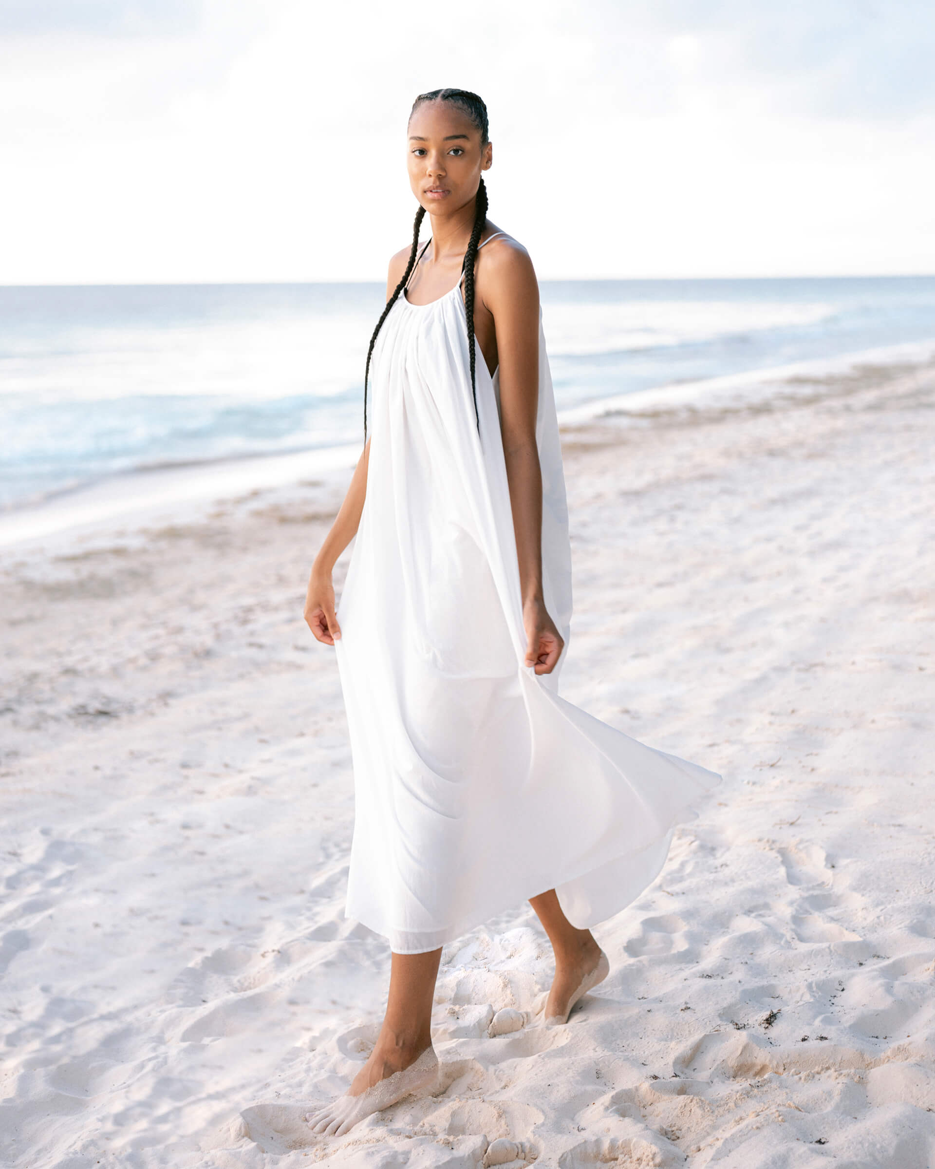 female wearing white maxi dress holding the dress with both hands and walking on the beach 