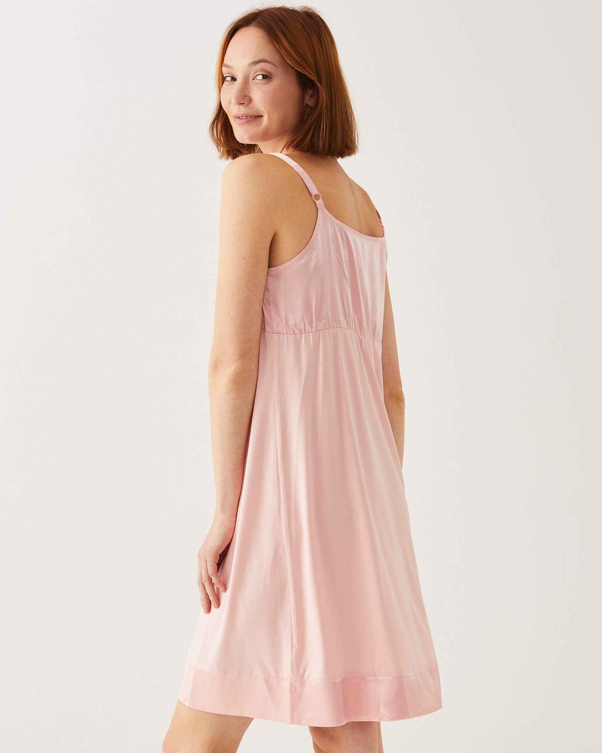 female wearing light pink satin pajama sleep dress looking over a shoulder on a white background