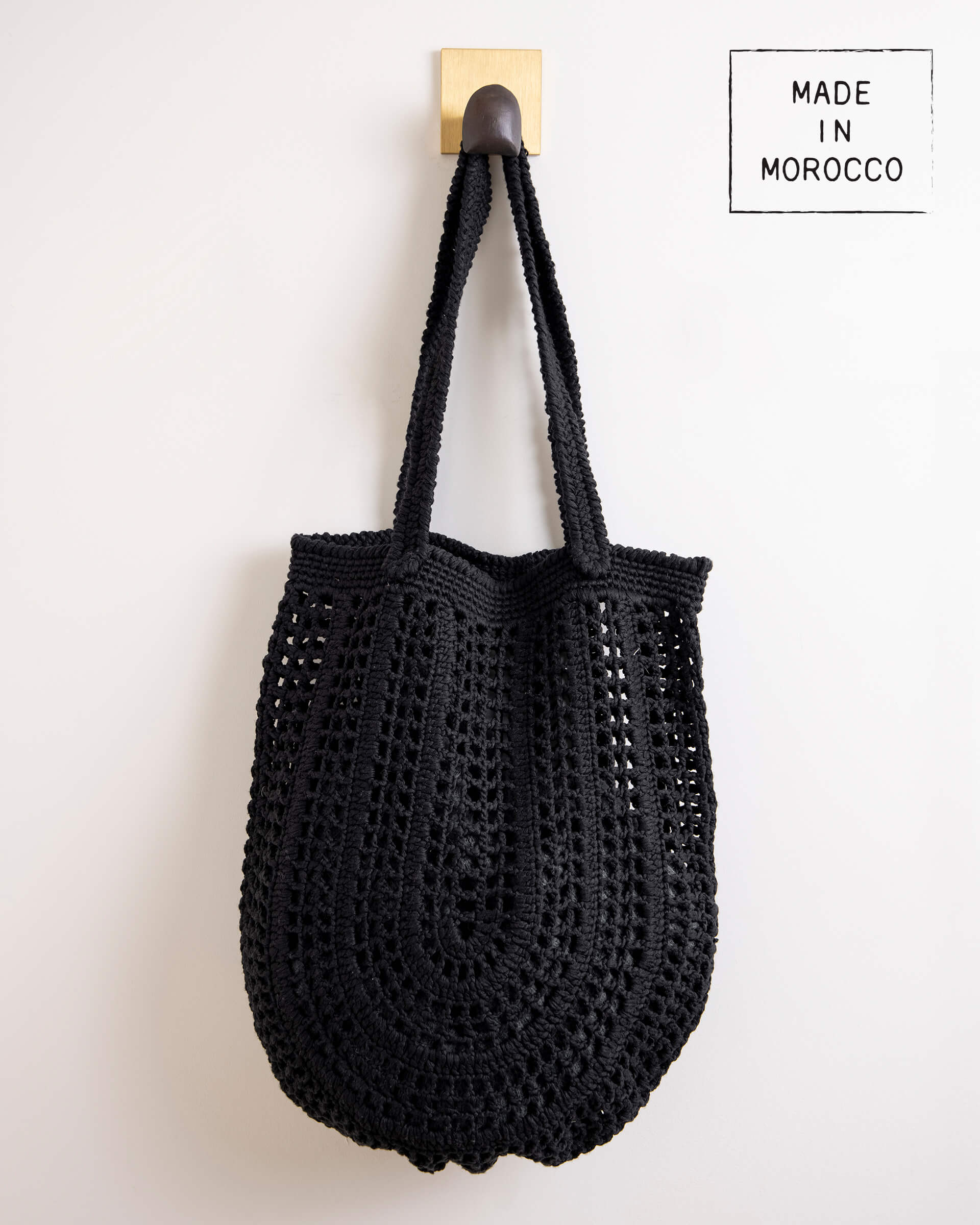 Souk Crochet Tote - Blackblack crochet tote with a long strap and several holes hanging on a hook on a white background