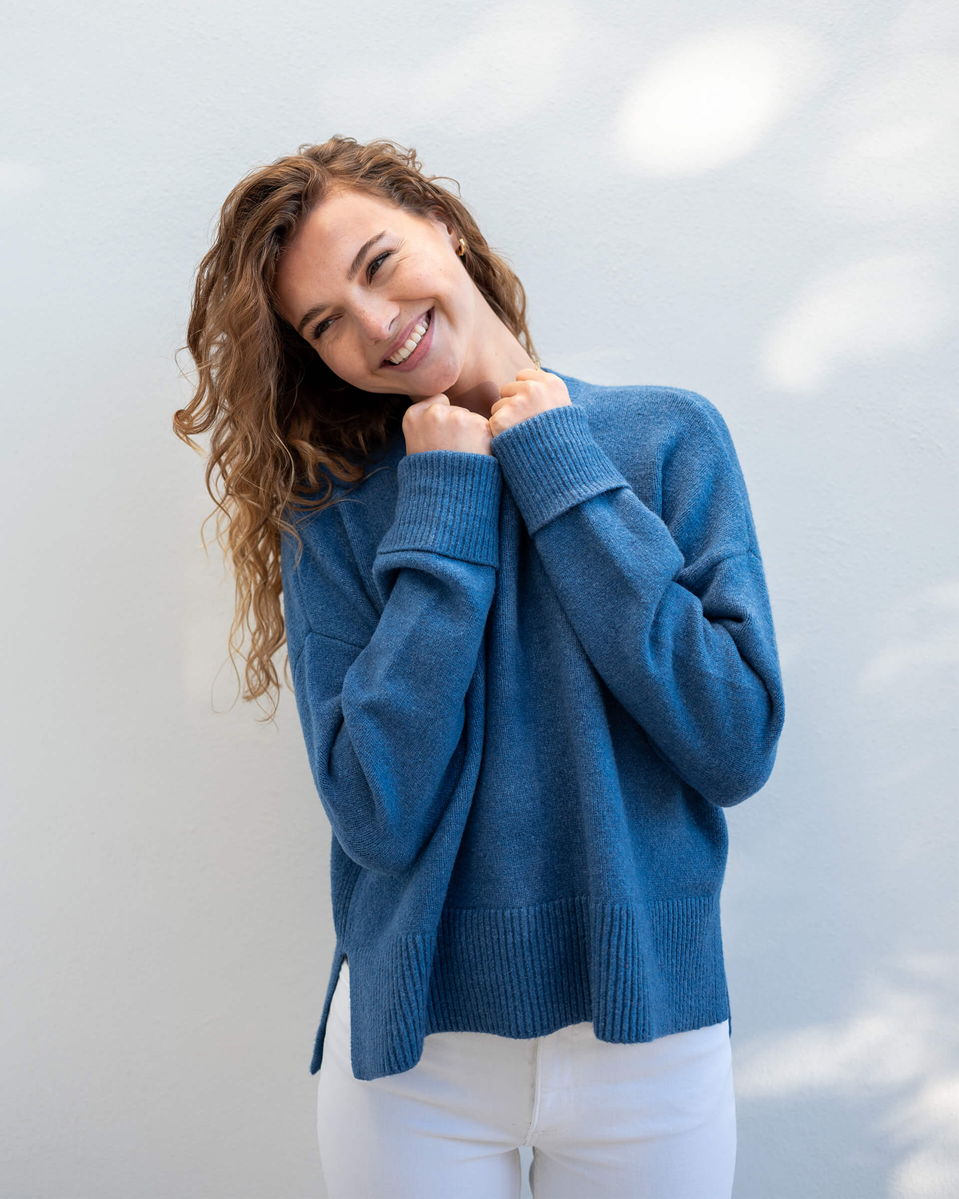female wearing denim blue crewneck sweater standing in front of a white wall 