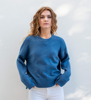female wearing denim blue crewneck sweater standing in front of a white wall with hands in pockets
