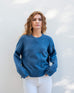 female wearing denim blue crewneck sweater standing in front of a white wall with hands in pockets