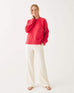 female wearing red crewneck sweater standing with an arm to her neck on a white background