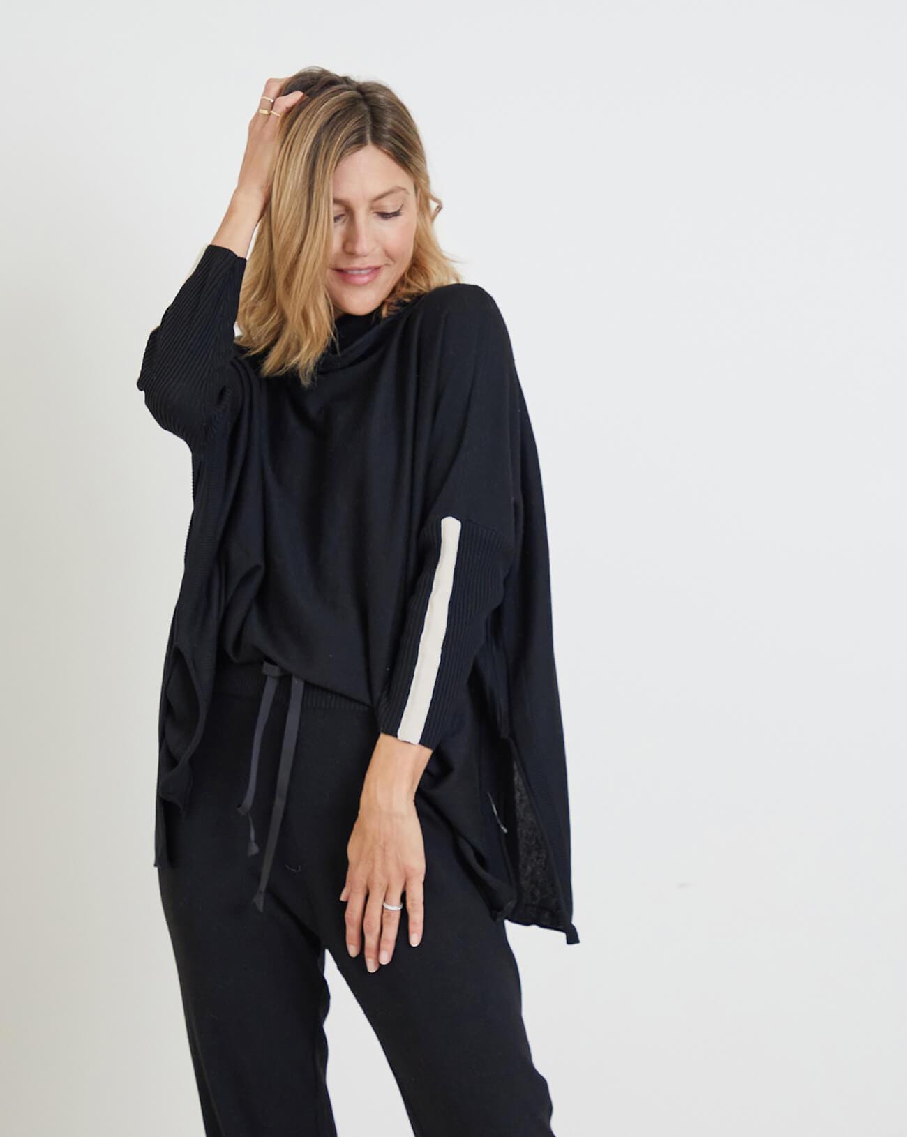woman wearing mersea newport sweater in black with white accent stripe down lower sleeve with a split front turtleneck and ribbed sleeves with right hand held up to head
