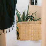 large straw basket wrapped with black leather handle sitting on a ledge outside near plants 