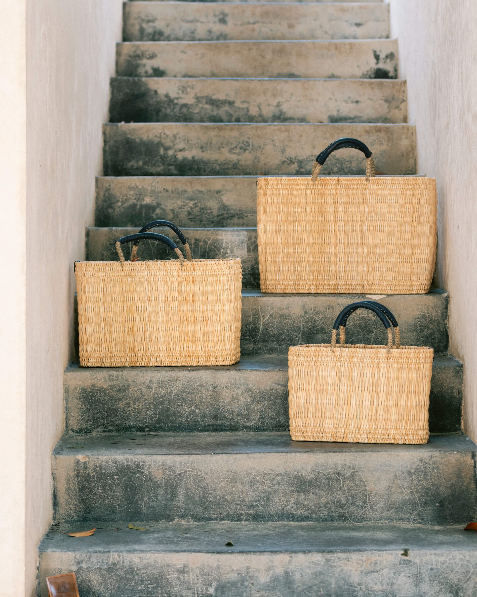 small, medium, large straw basket wrapped with black leather handles sitting on different stairs 