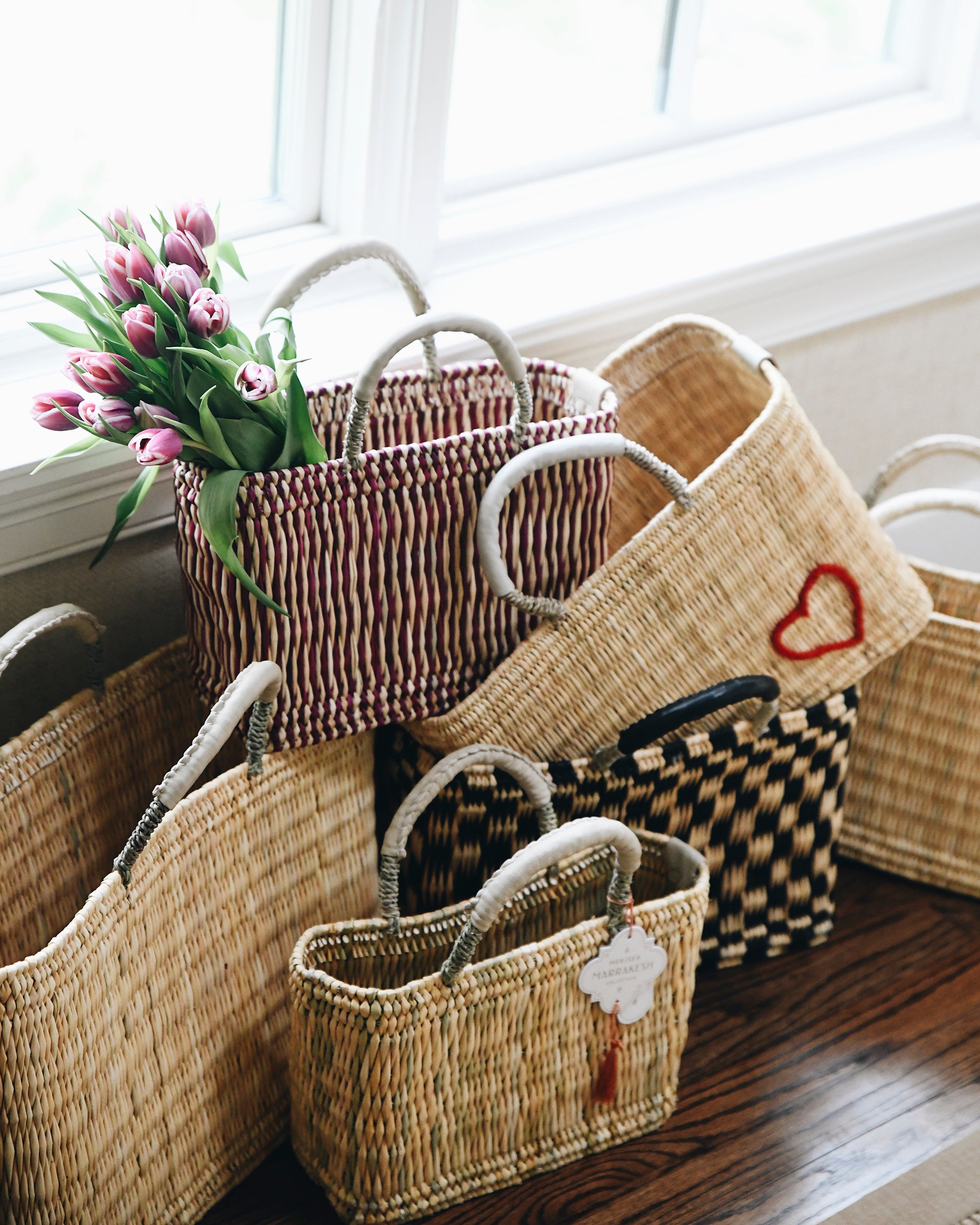six different colored and sized straw baskets stacked near a window on a hardwood floor