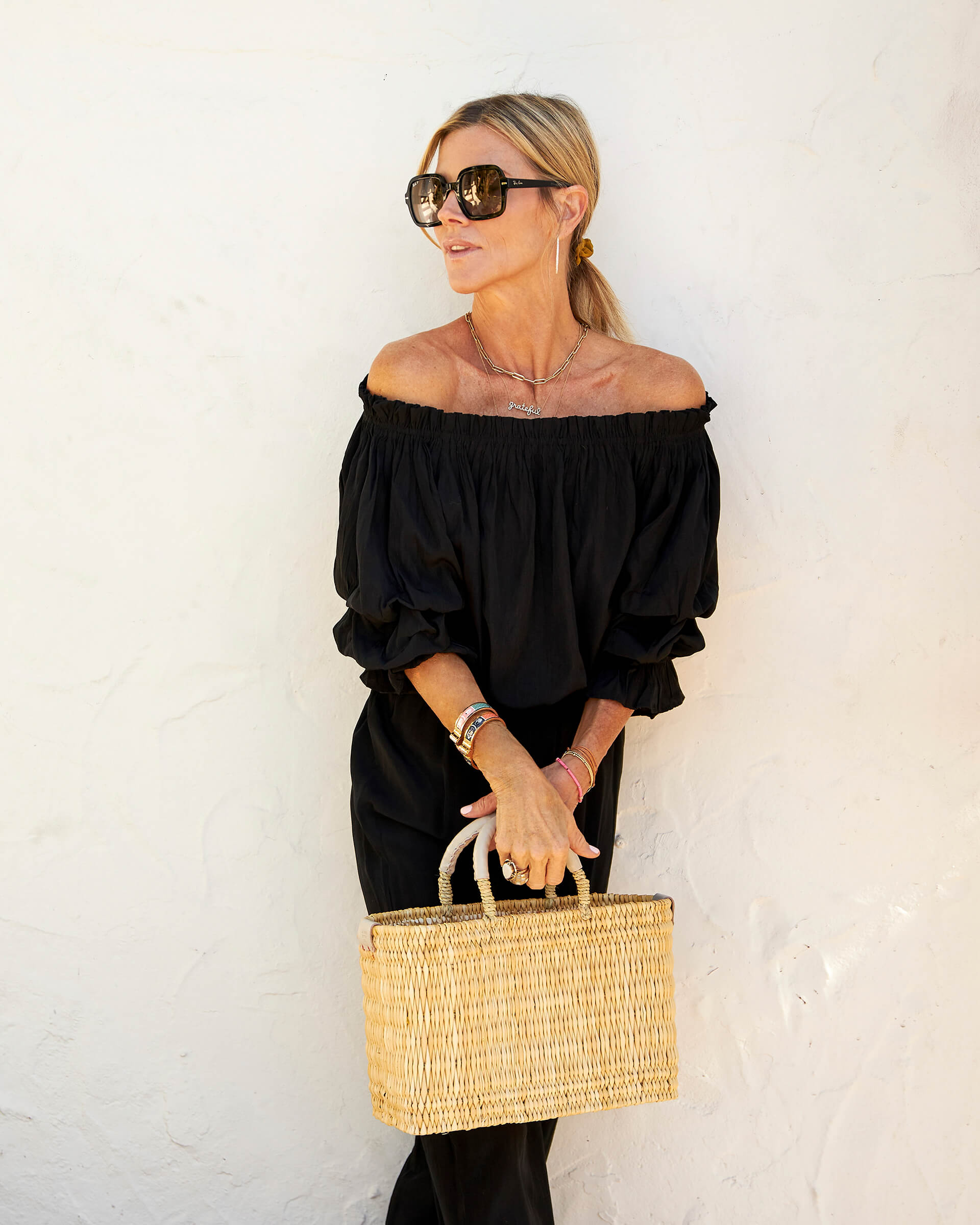 blonde female holding small straw basket with a neutral colored handle leaning against white wall