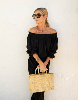 blonde female holding small straw basket with a neutral colored handle leaning against white wall