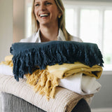 female holding a stack of grey, light beige, yellow, white, and dark blue gauze and shibori blankets
