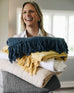 female holding a stack of grey, light beige, yellow, white, and dark blue gauze and shibori blankets