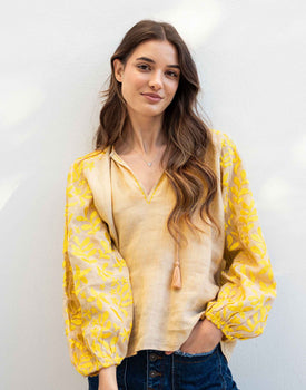 female wearing tan and yellow embroidered blouse with tassel ties standing in front of a white wall