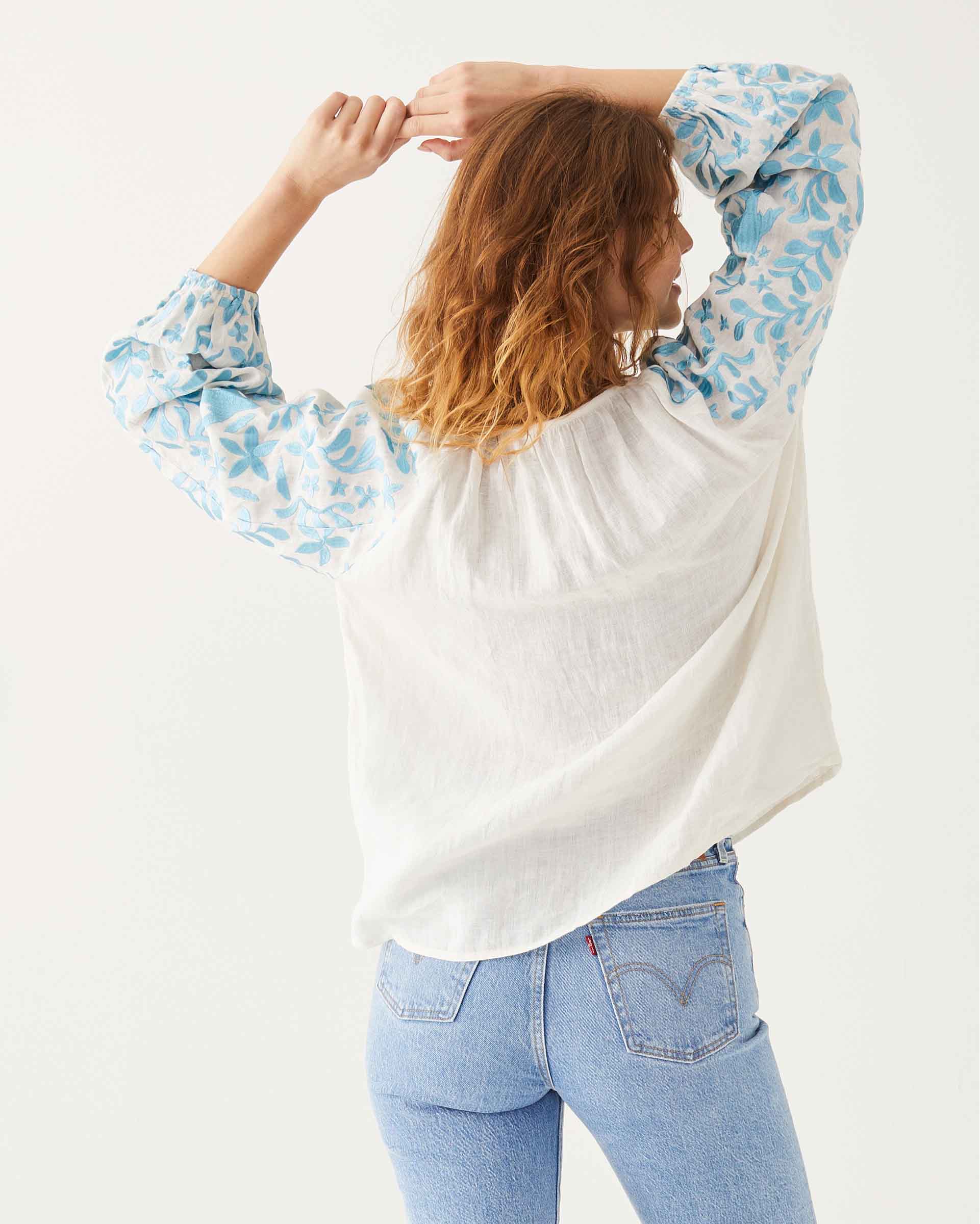 female wearing white and blue embroidered blouse with vines and birds backwards on white background