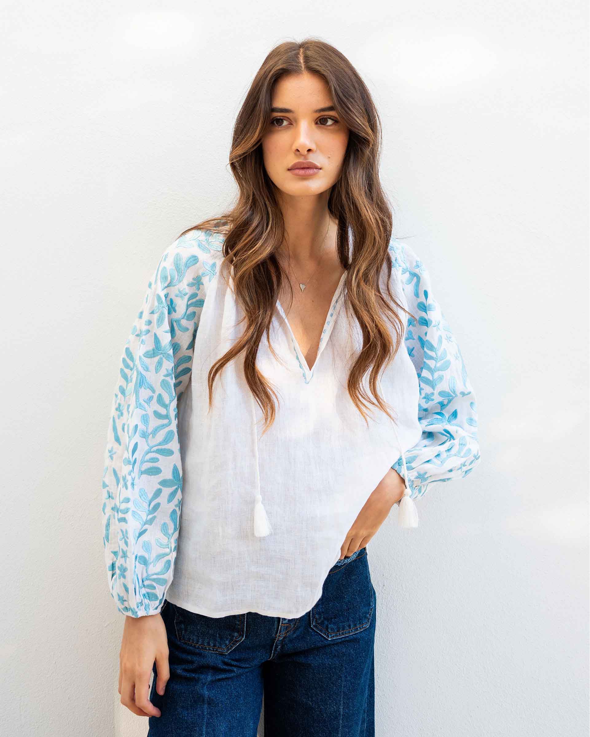 female wearing white and blue embroidered blouse with v-neckline standing in front of a white wall
