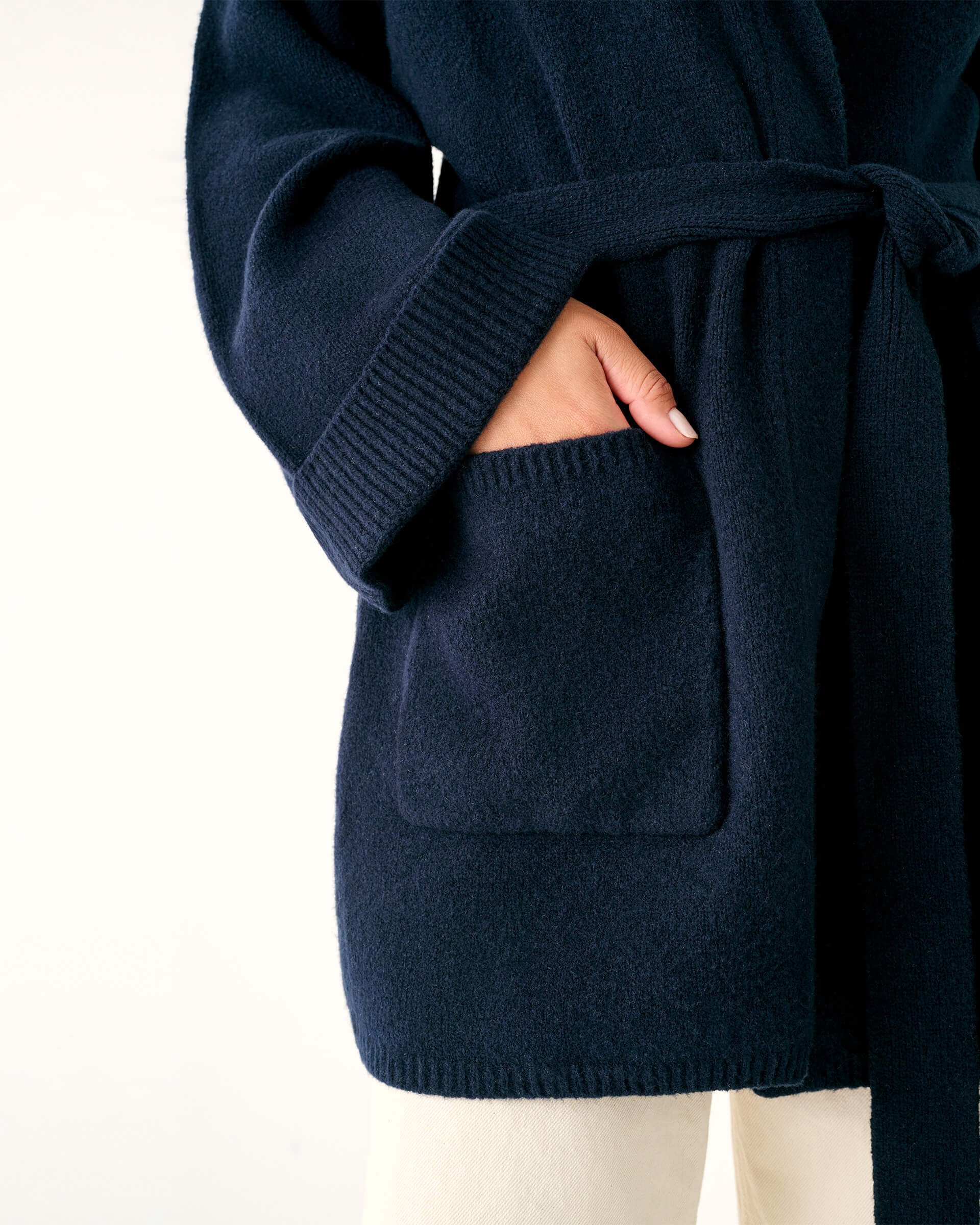 close up of female wearing navy wrap sweater with hand in pocket and belt on a white background 
