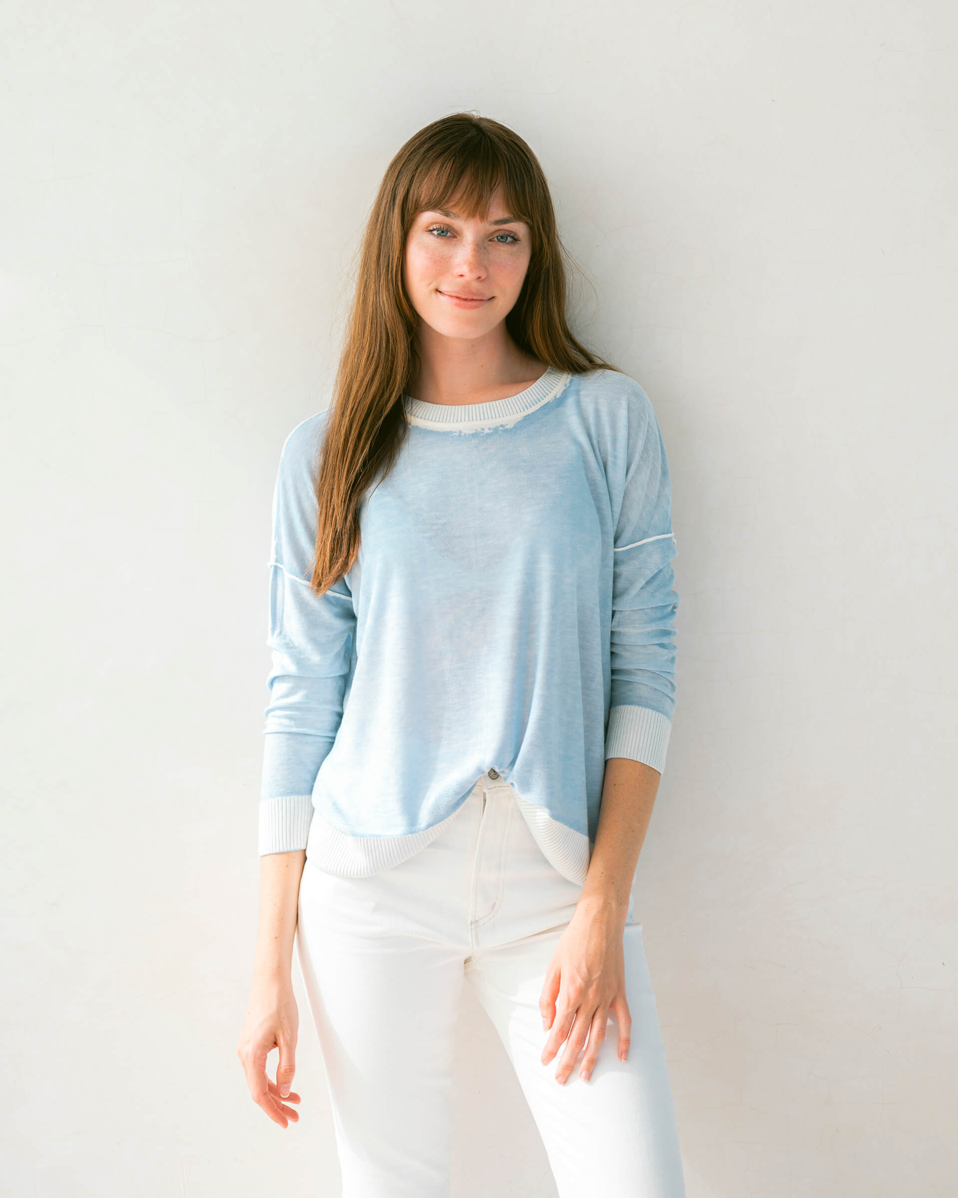 brunette female wearing light blue sweater standing in front of a white wall