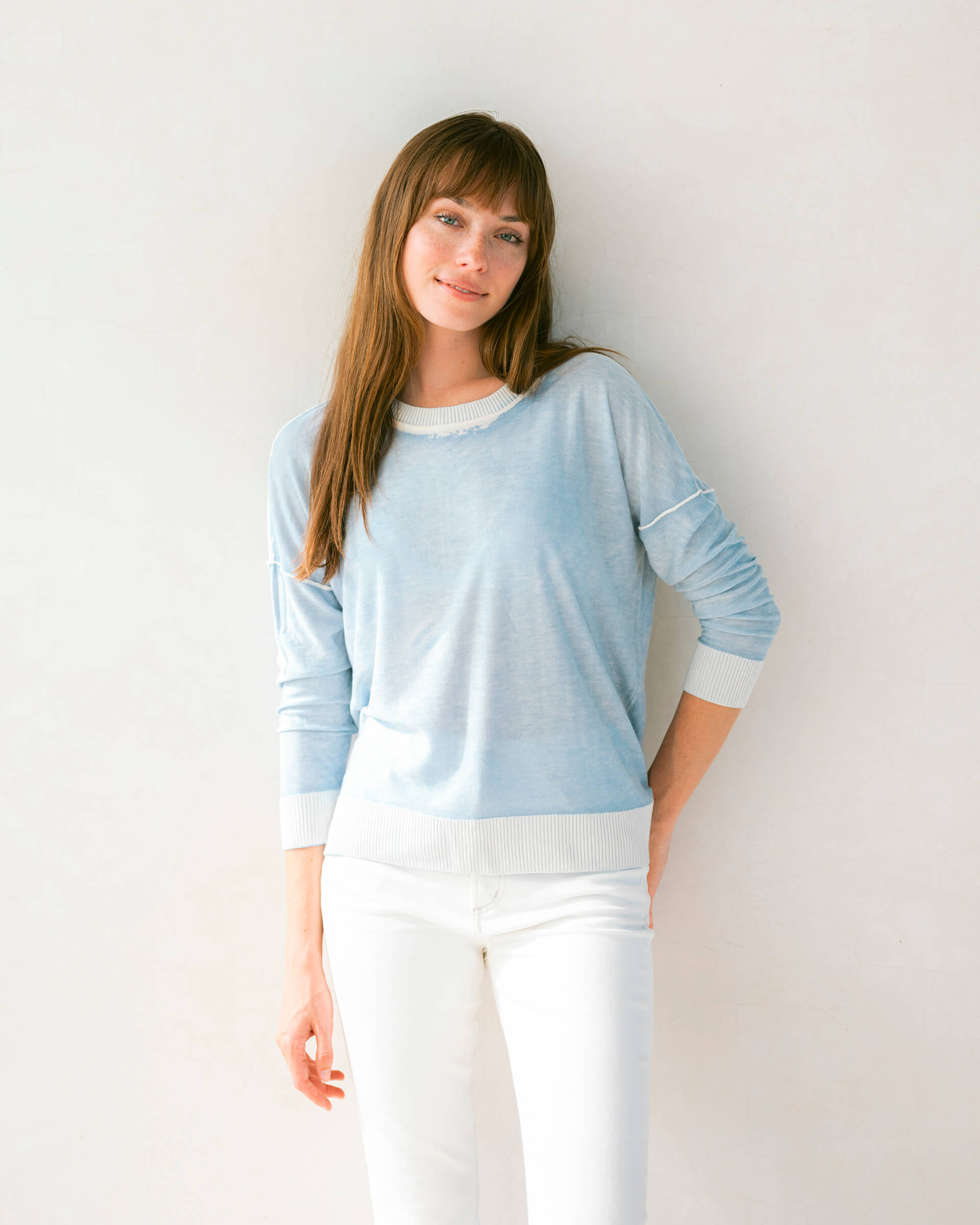 brunette female wearing light blue sweater standing in front of a white wall with an arm on her hip