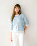 brunette female wearing light blue sweater standing in front of a white wall with an arm on her hip