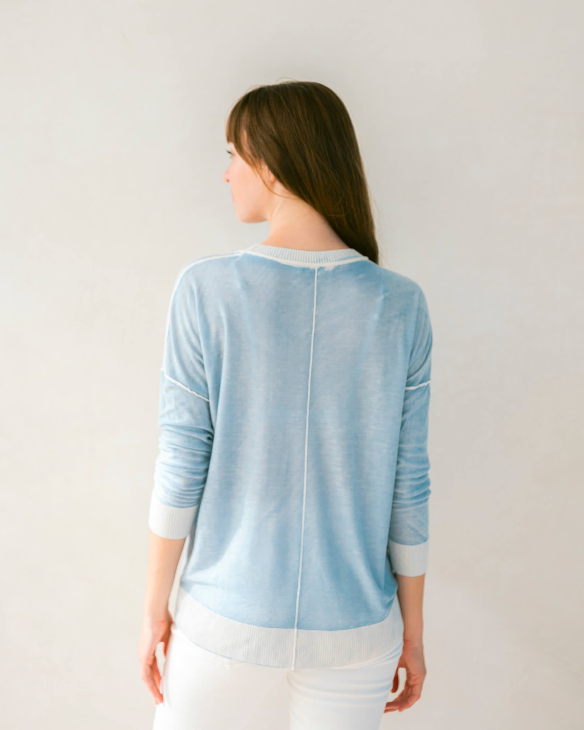 brunette female wearing light blue sweater standing backwards in front of a white wall 