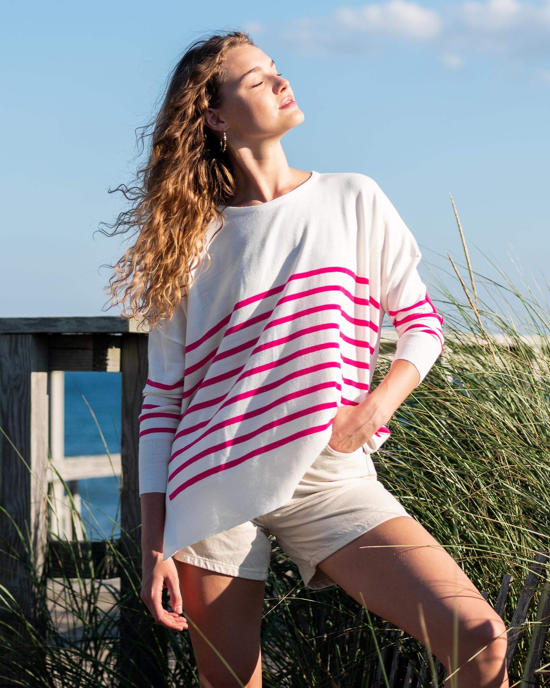 female wearing white and pink striped sweater standing looking at the sun on the beach 