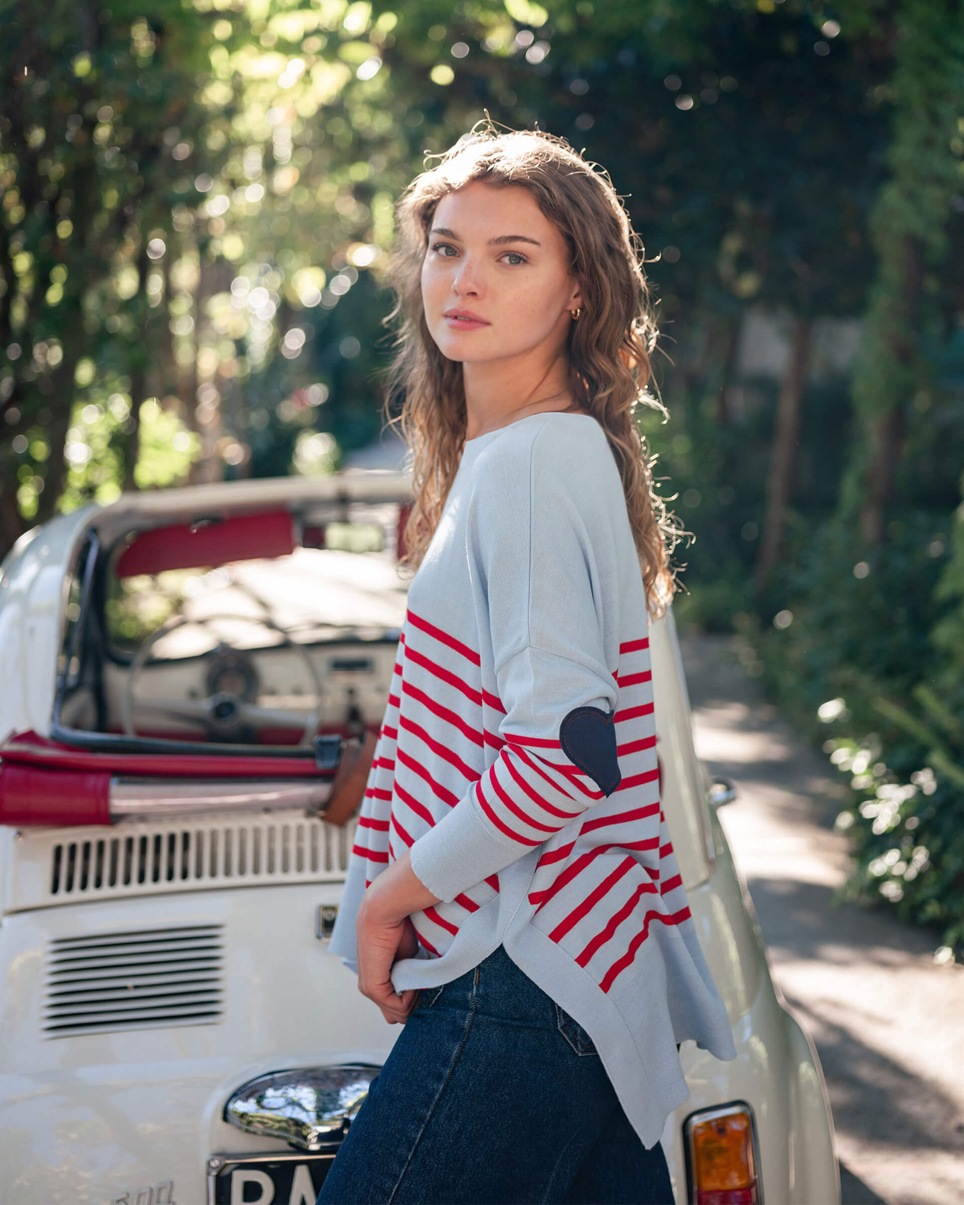 female wearing light blue and red striped sweater standing sideways in front of a white car outside 