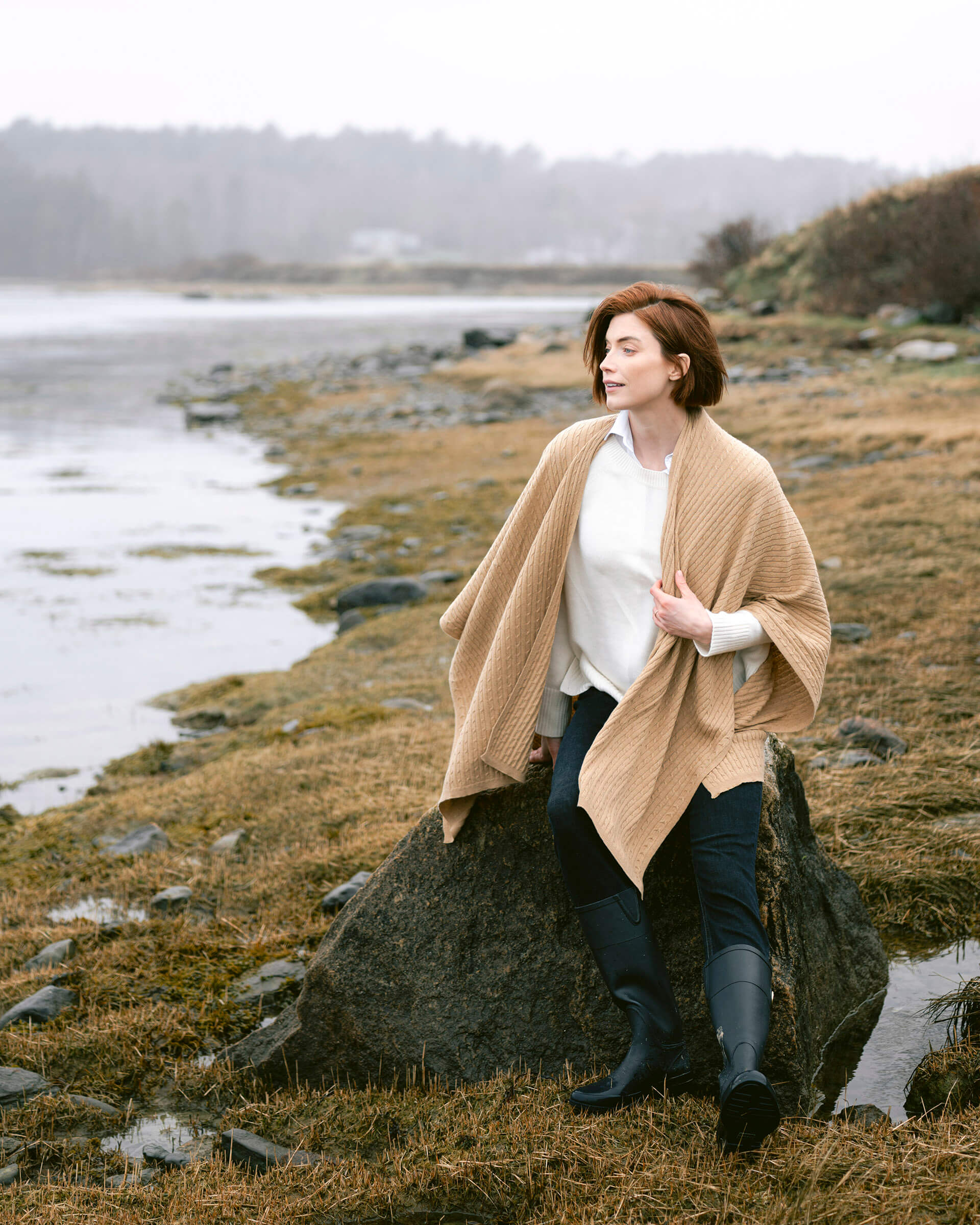 female wearing golden brown cable knit wrap sitting on a rock near the sea coast