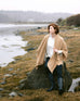 female wearing golden brown cable knit wrap sitting on a rock near the sea coast
