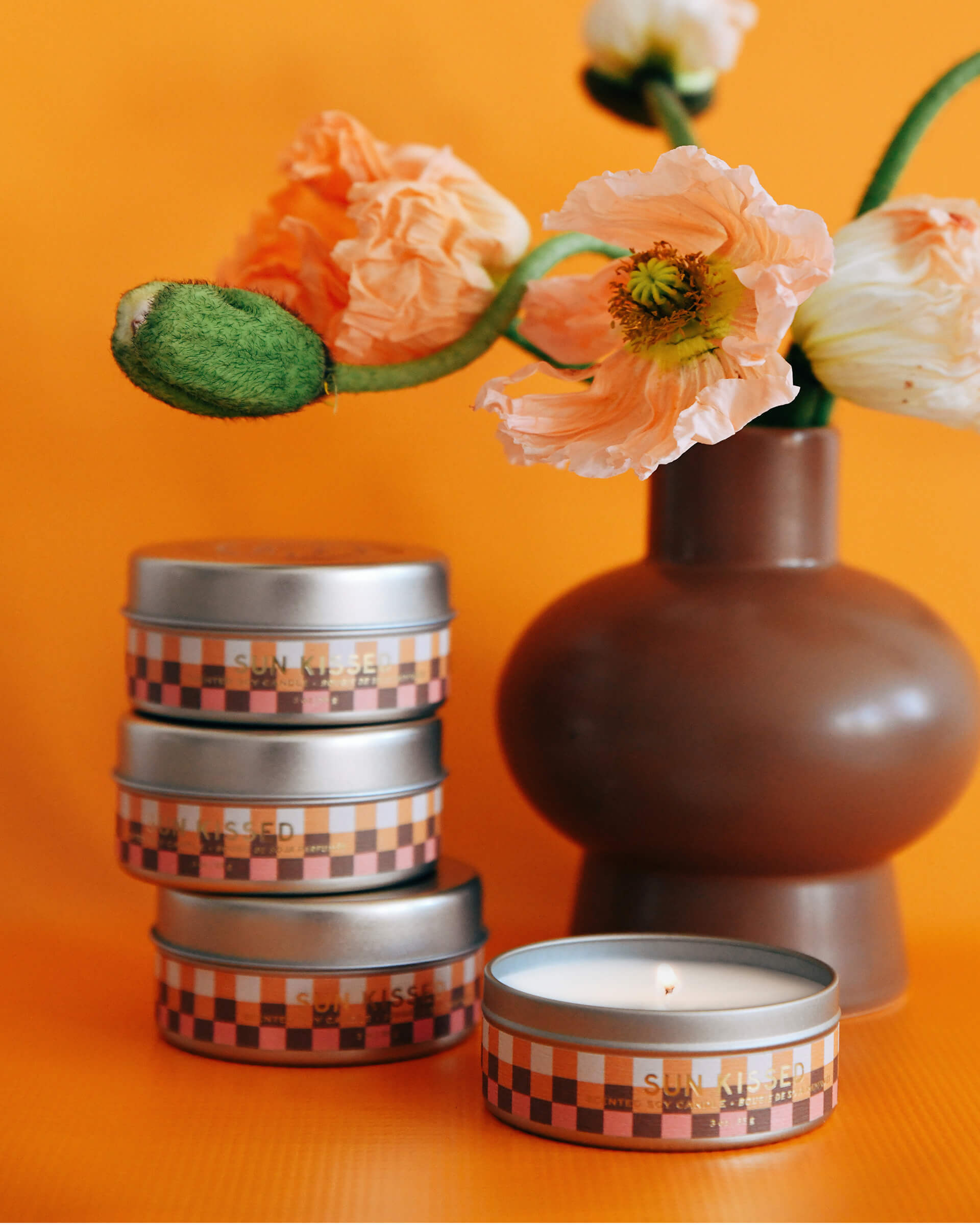 four orange and white checkered 3 oz tin sun kissed candles with flower vase on orange background