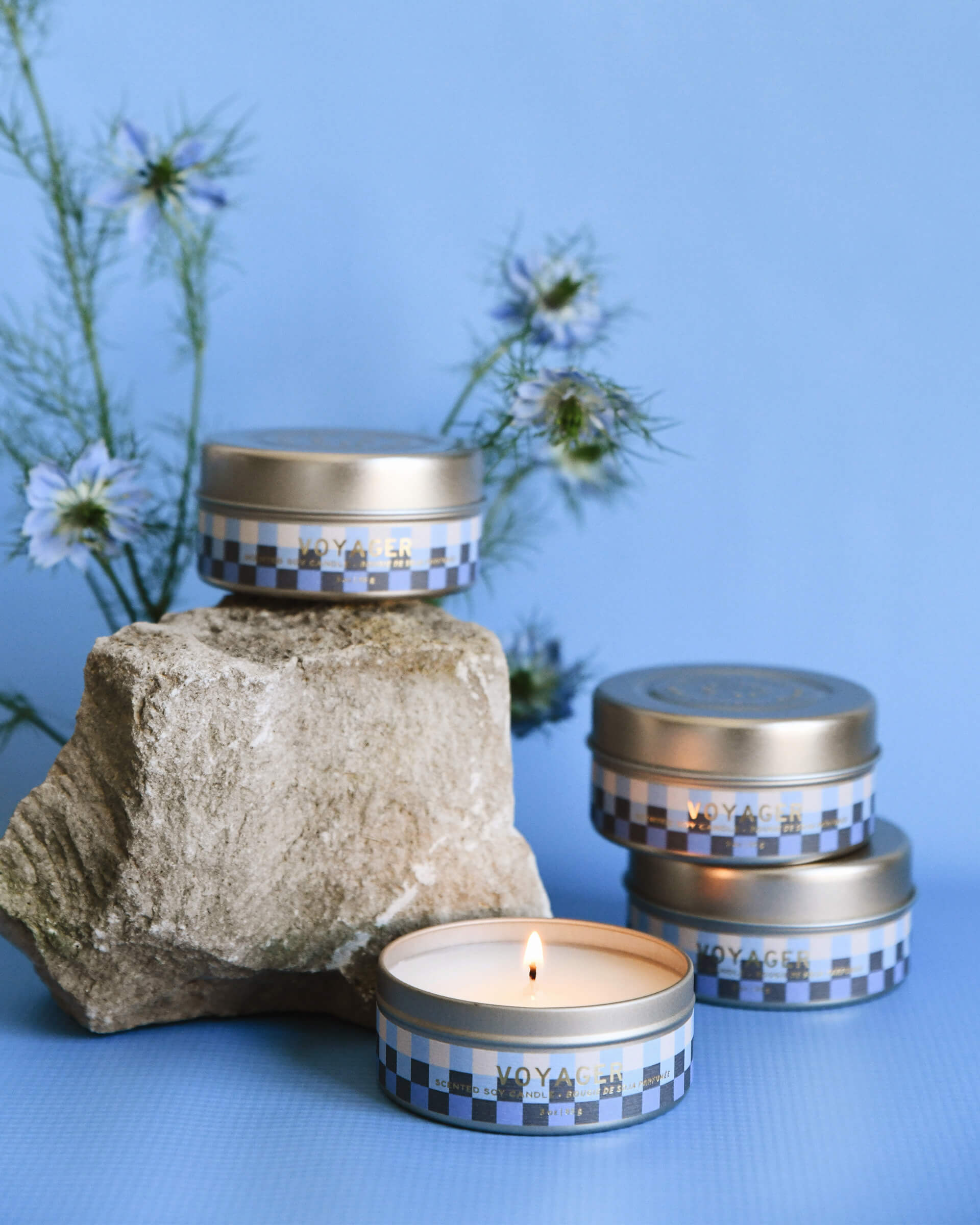 four blue and white checkered 3 oz tin voyager candles sitting near a rock on a blue background