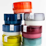 seven diferent colored canister candles stacked on top of one another on a white background