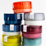 seven diferent colored canister candles stacked on top of one another on a white background