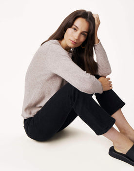 brunette female wearing light brown sweater sitting down with her knees up on a white background 