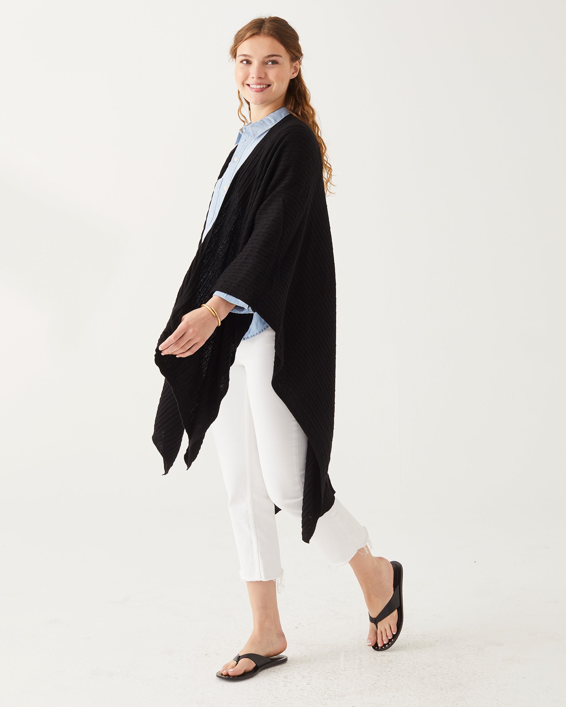 Female wearing a black wrap over a blue button shirt and white jeans standing in front of white wall