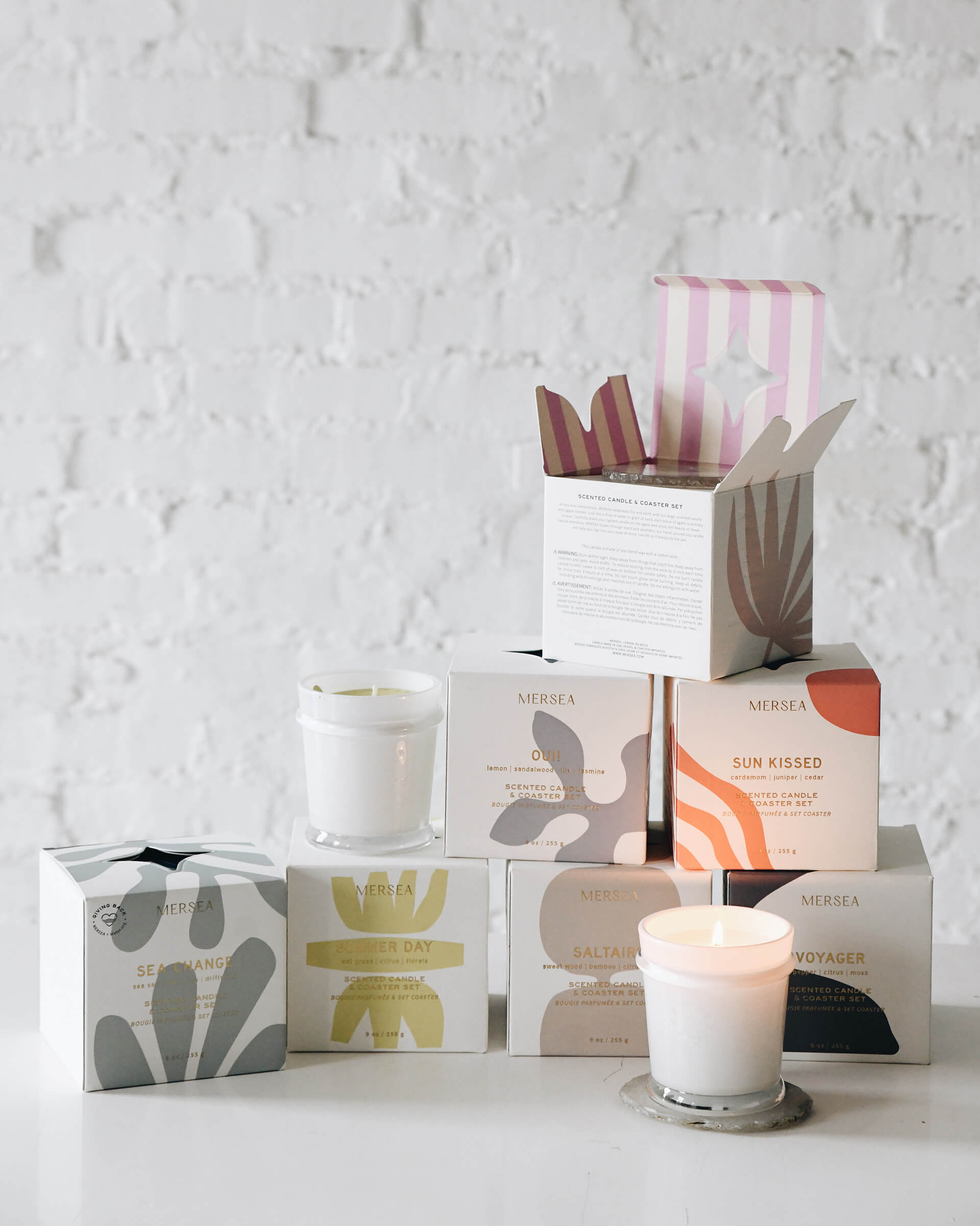 collection of several boxed candles with coasters and two unboxed white candles on a white wall