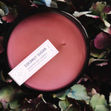 7 oz dark pink coconut sugar canister candle surrounded by green and dark pink leaves 