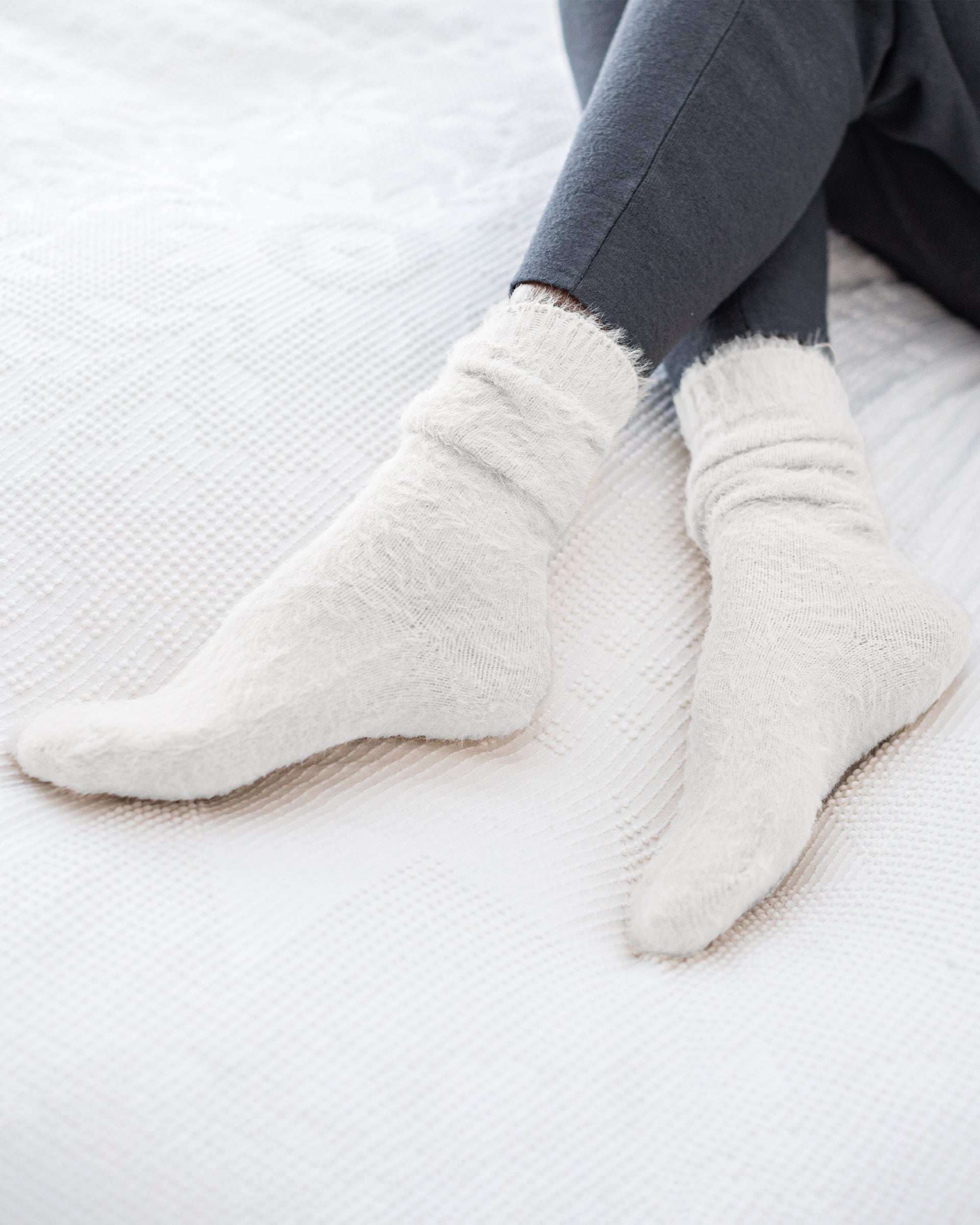 female wearing white fuzzy socks sitting on a bed with her legs crossed on a white comforter 