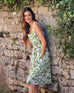 female wearing white and green floral A-line linen sundress with self-belt leaning on a stone wall 