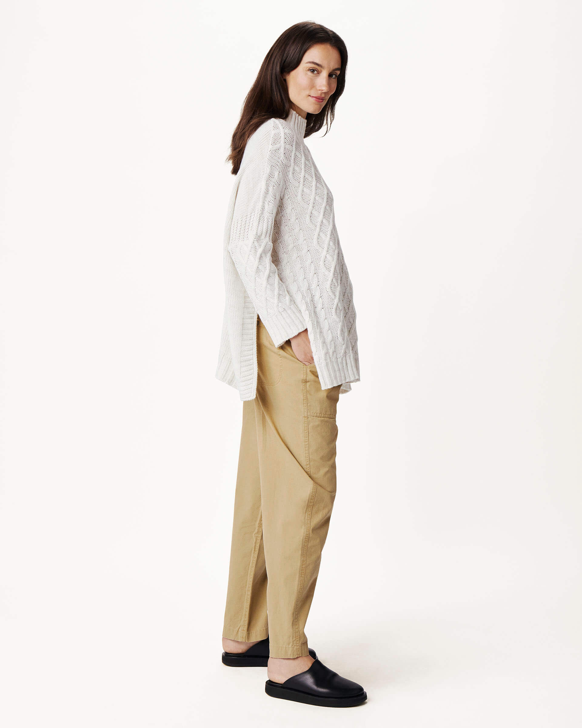 brunette female wearing white sweater turned sideways with hand in pocket on a white background