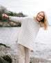 blonde female wearing white sweater standing on rocks on a beach smiling with an arm raised